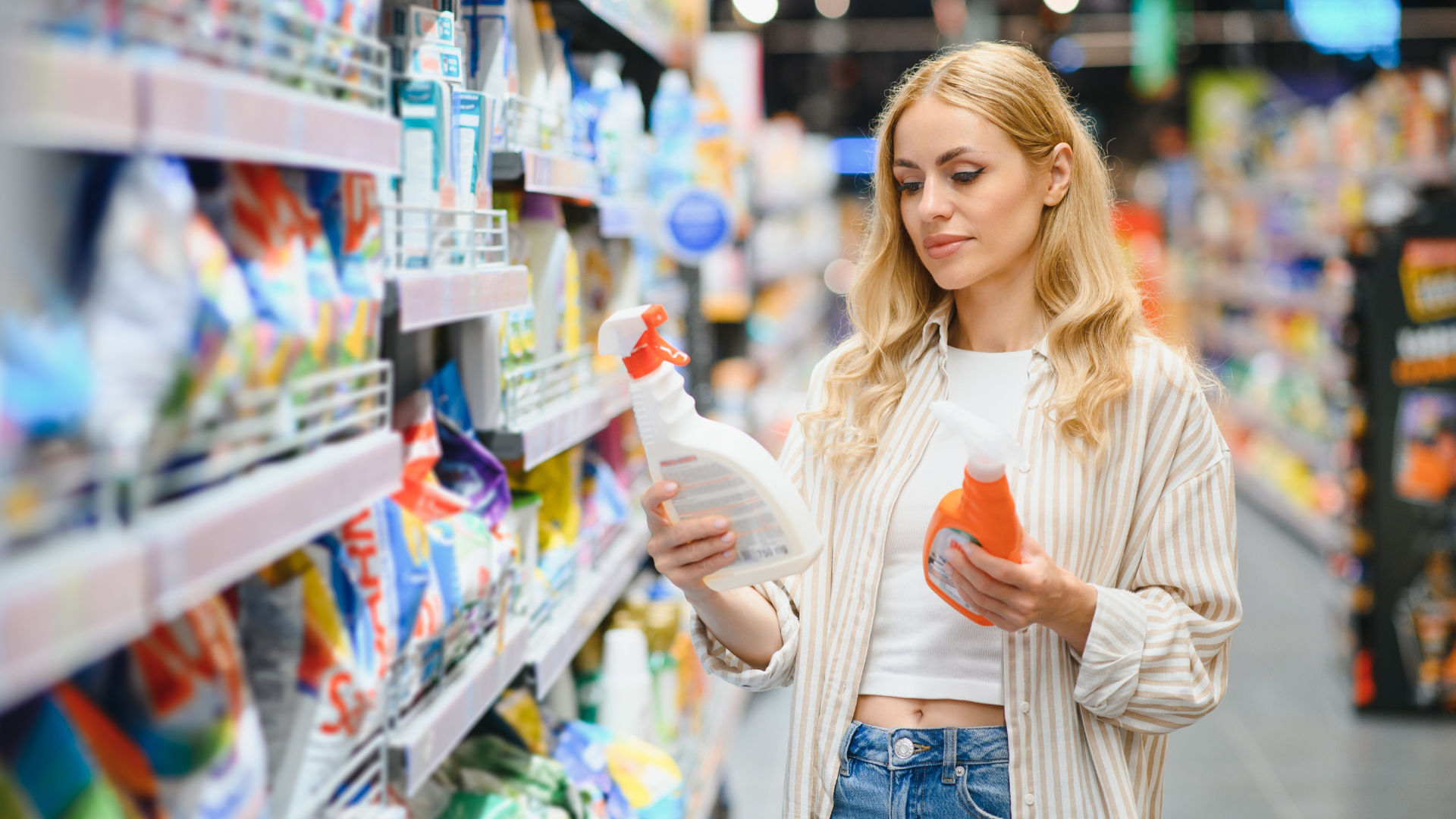 Una mujer que está comprando en el pasillo de una tienda de comestibles, comparando dos productos de limpieza.
