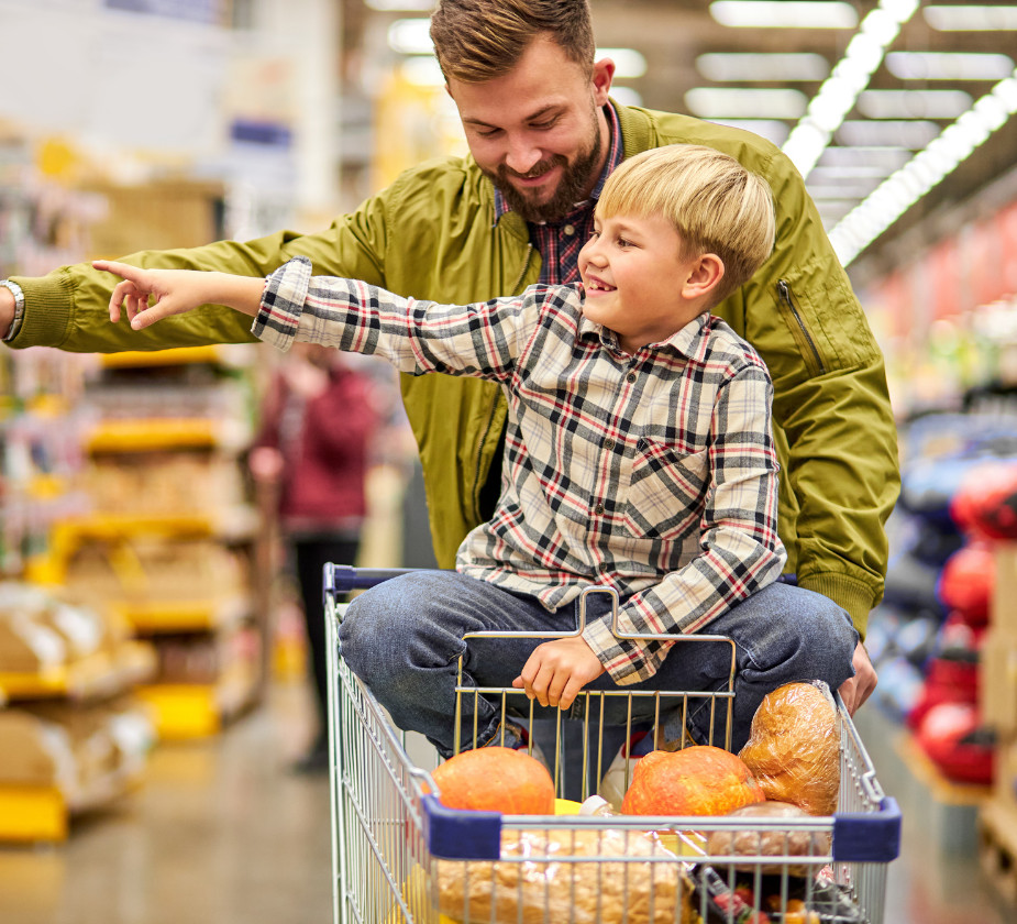 A man smiling while pushing a shopping cart with a young boy sitting inside, pointing in a grocery store.