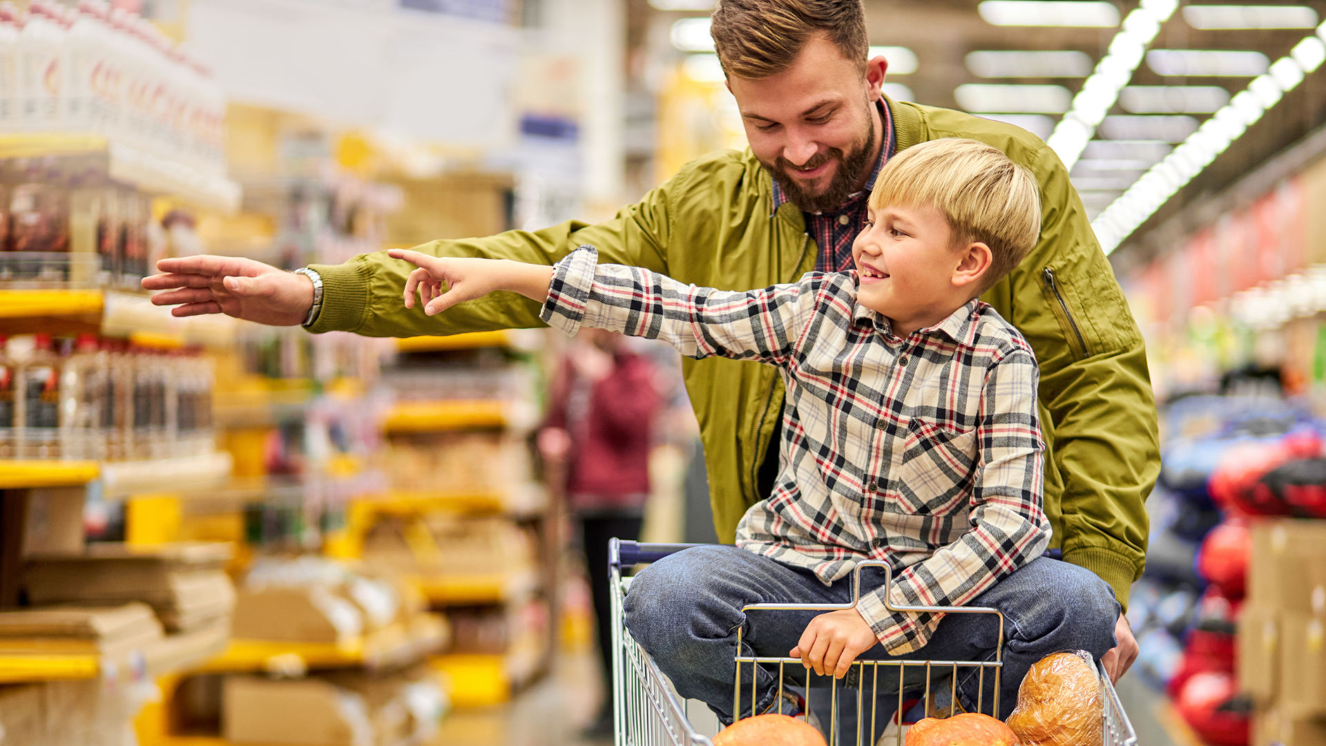 A man smiling while pushing a shopping cart with a young boy sitting inside, pointing in a grocery store.