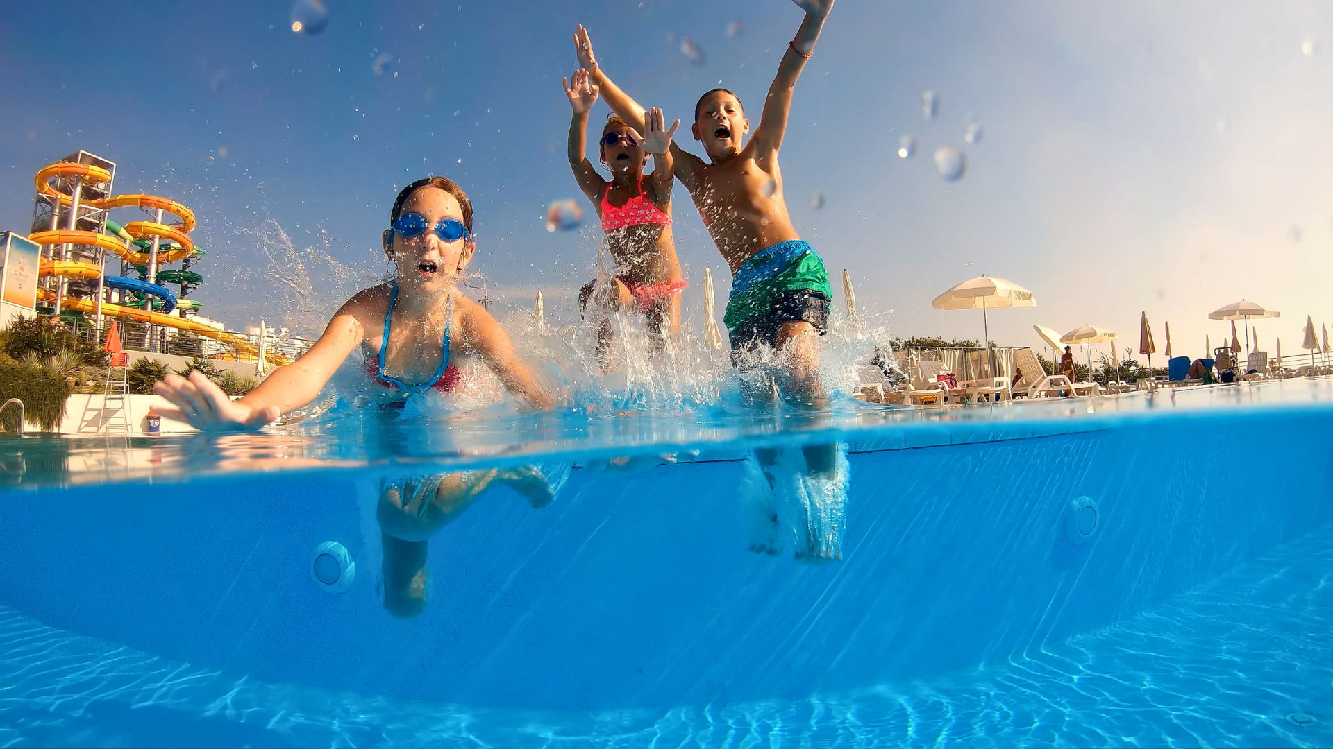 A group of children jumping into a swimming pool, creating splashes, with water slides and sunny skies in the background.