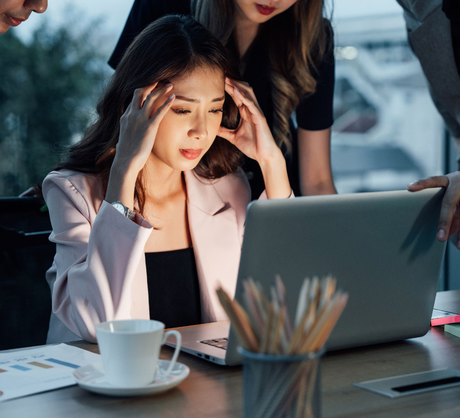 A woman is sitting at a table looking stressed while working on a laptop. Blurry figures are walking in the background
