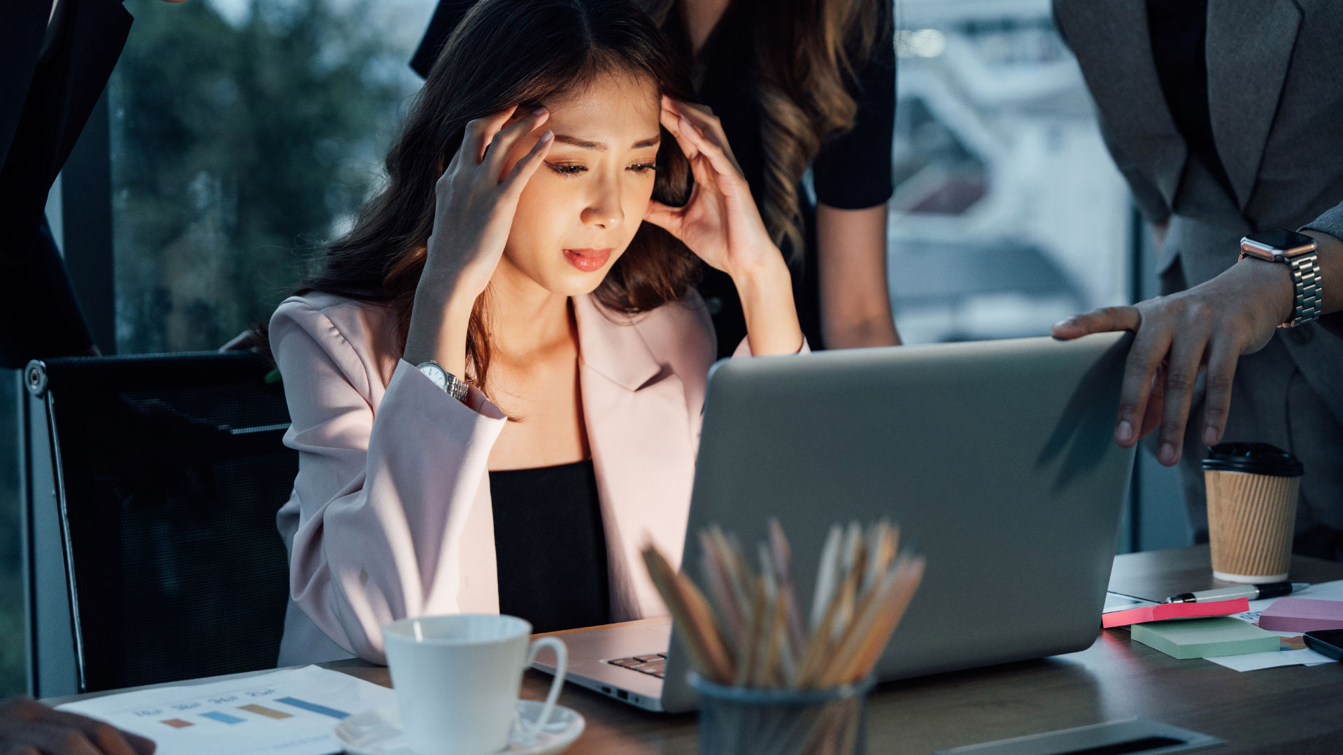 A woman is sitting at a table looking stressed while working on a laptop. Blurry figures are walking in the background