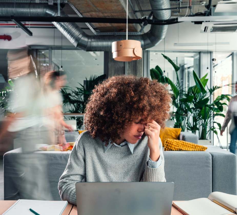 A woman is sitting at a table looking stressed while working on a laptop. Blurry figures are walking in the background
