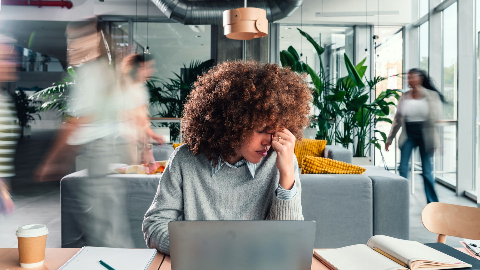 A woman is sitting at a table looking stressed while working on a laptop. Blurry figures are walking in the background