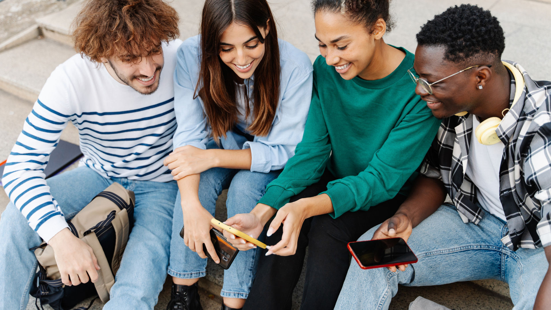 A group of four friends sitting together outdoors, looking at a smartphone and smiling.