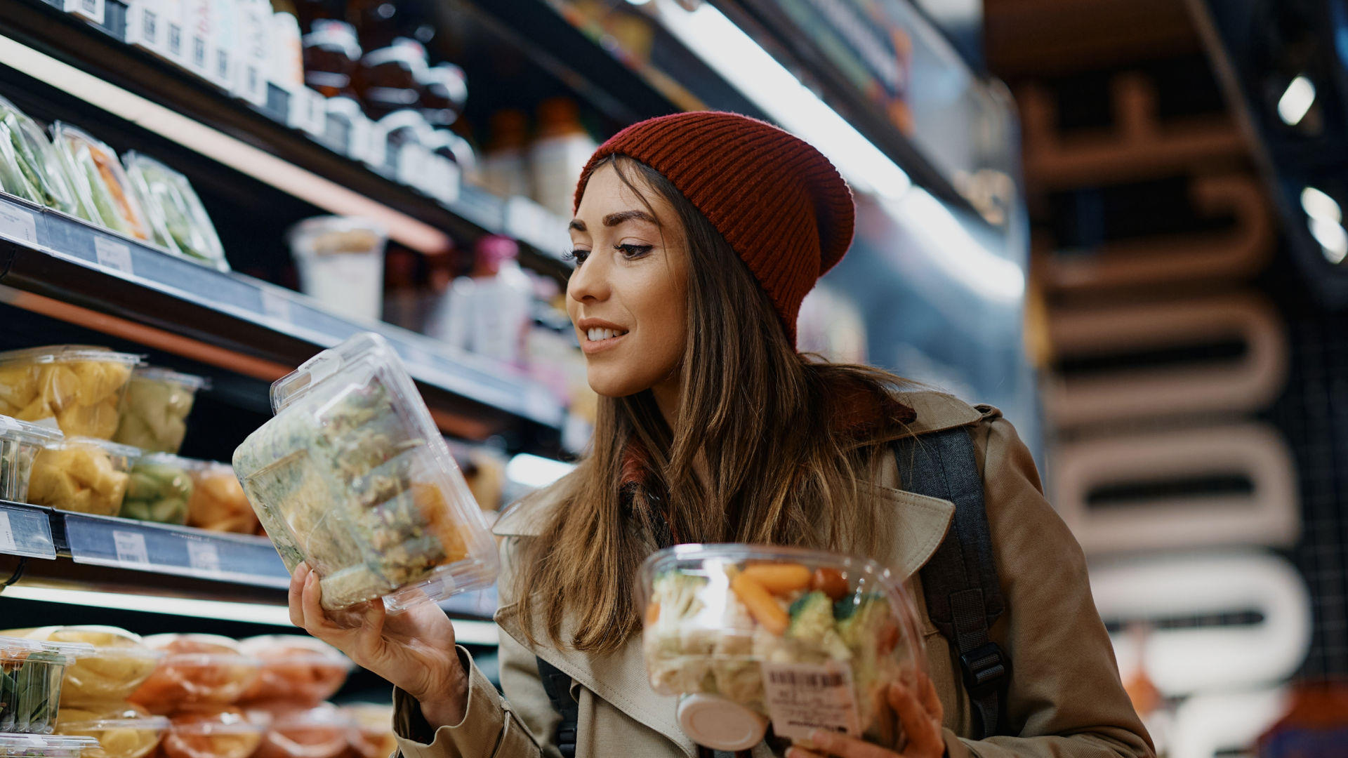 A woman shopping in a grocery store, holding two plastic containers of pre-packaged fruits and vegetables, with shelves of produce in the background.