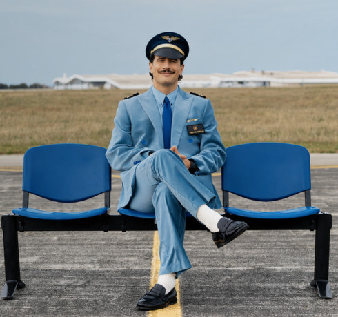 A man in an airline uniform and pilot's cap sitting on a blue bench at an airport.