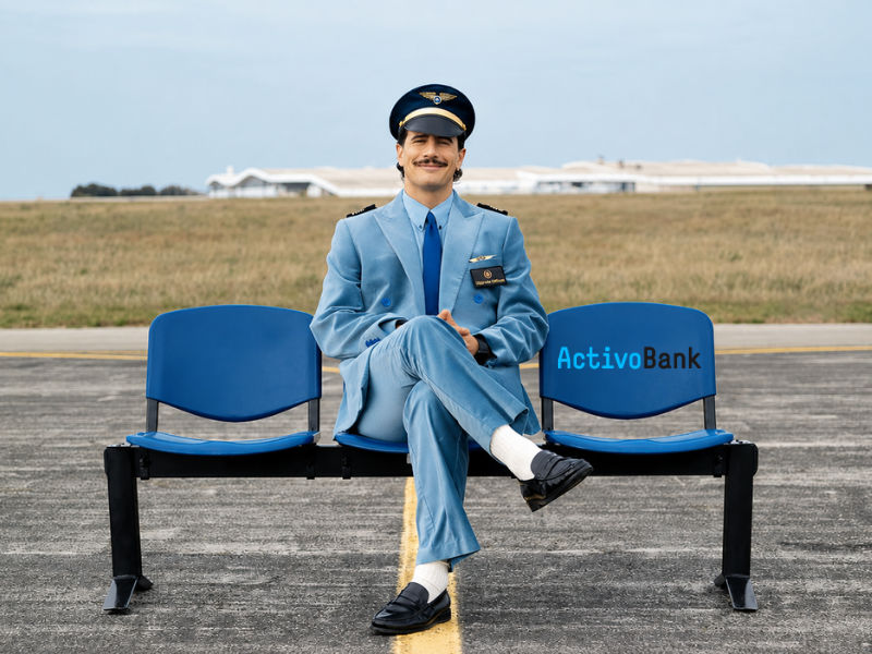 A man in an airline uniform and pilot's cap sitting on a blue bench at an airport.