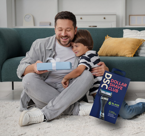 A man and a boy sitting on the floor, smiling and hugging, with a gift box and a packaging of Dollar Shave Club razors