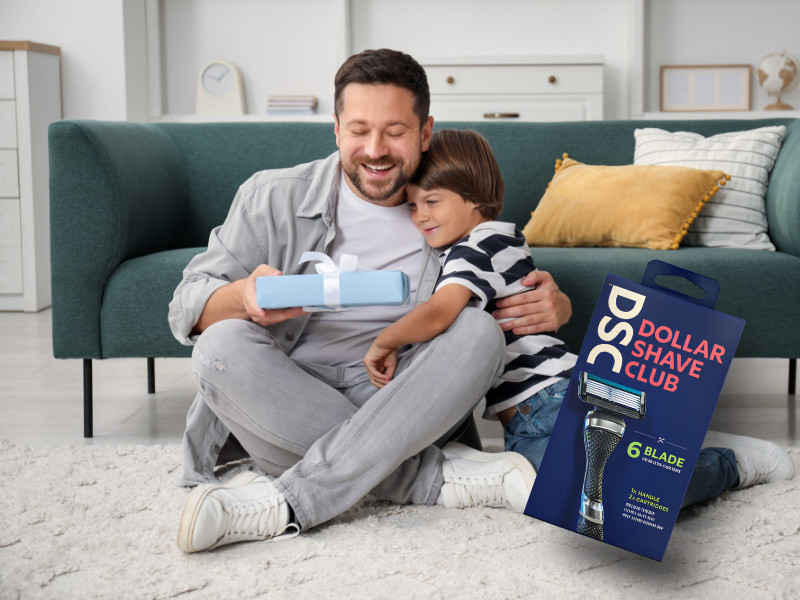 A man and a boy sitting on the floor, smiling and hugging, with a gift box and a packaging of Dollar Shave Club razors