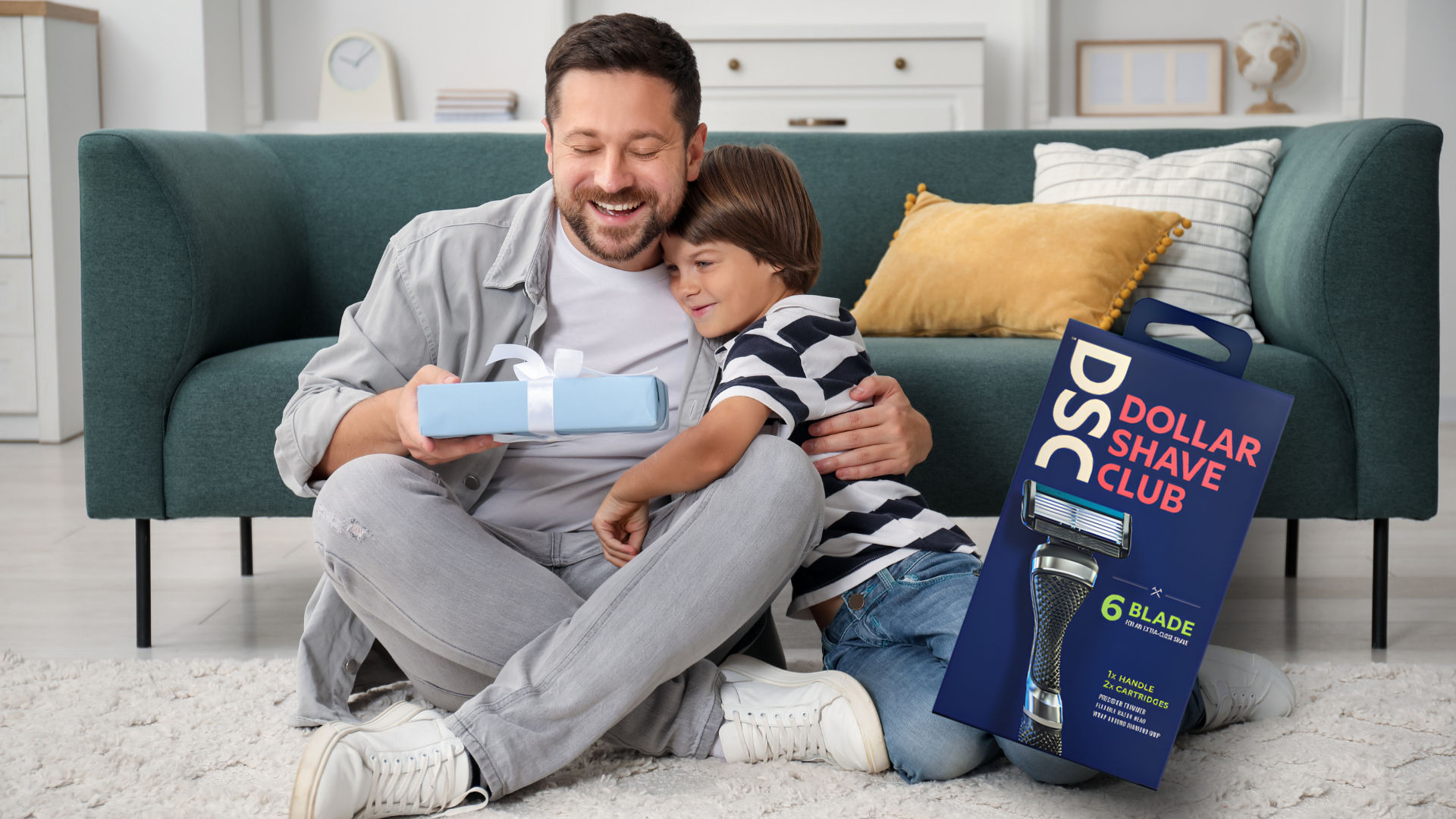 A man and a boy sitting on the floor, smiling and hugging, with a gift box and a packaging of Dollar Shave Club razors 