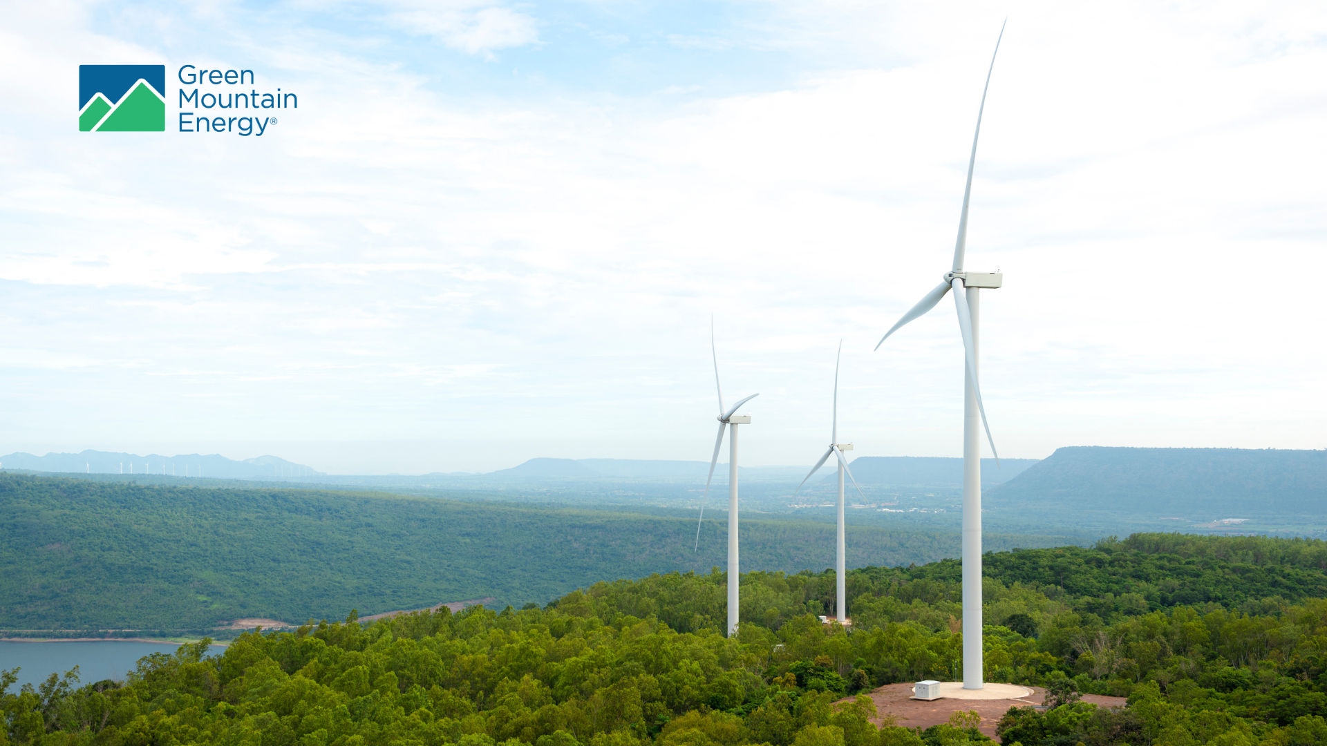Wind turbines overlooking a green forest and lake under a blue sky, representing renewable energy.
