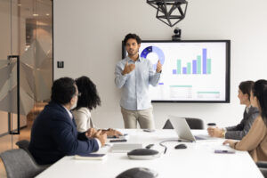 Junge männliche Fachkraft steht vor einem digitalen Bildschirm und präsentiert einer Gruppe von Kollegen, die um einen Konferenztisch herum sitzen, Geschäftsdaten in Balken- und Kreisdiagrammen. Strategieentwicklung, Analytik