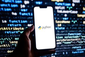 Auf einem Smartphone ist das Python-Logo zu erkennen. Das Smartphone wird von einer Hand vor einem Monitor gehalten, auf dem Python Codezeilen zu sehen sind.