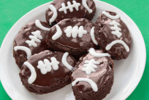 Homemade football shaped double chocolate fudge brownies with chocolate glazed icing and white icing to decorate like a football. The six brownies are served on a white plate and the background is green textured paper.