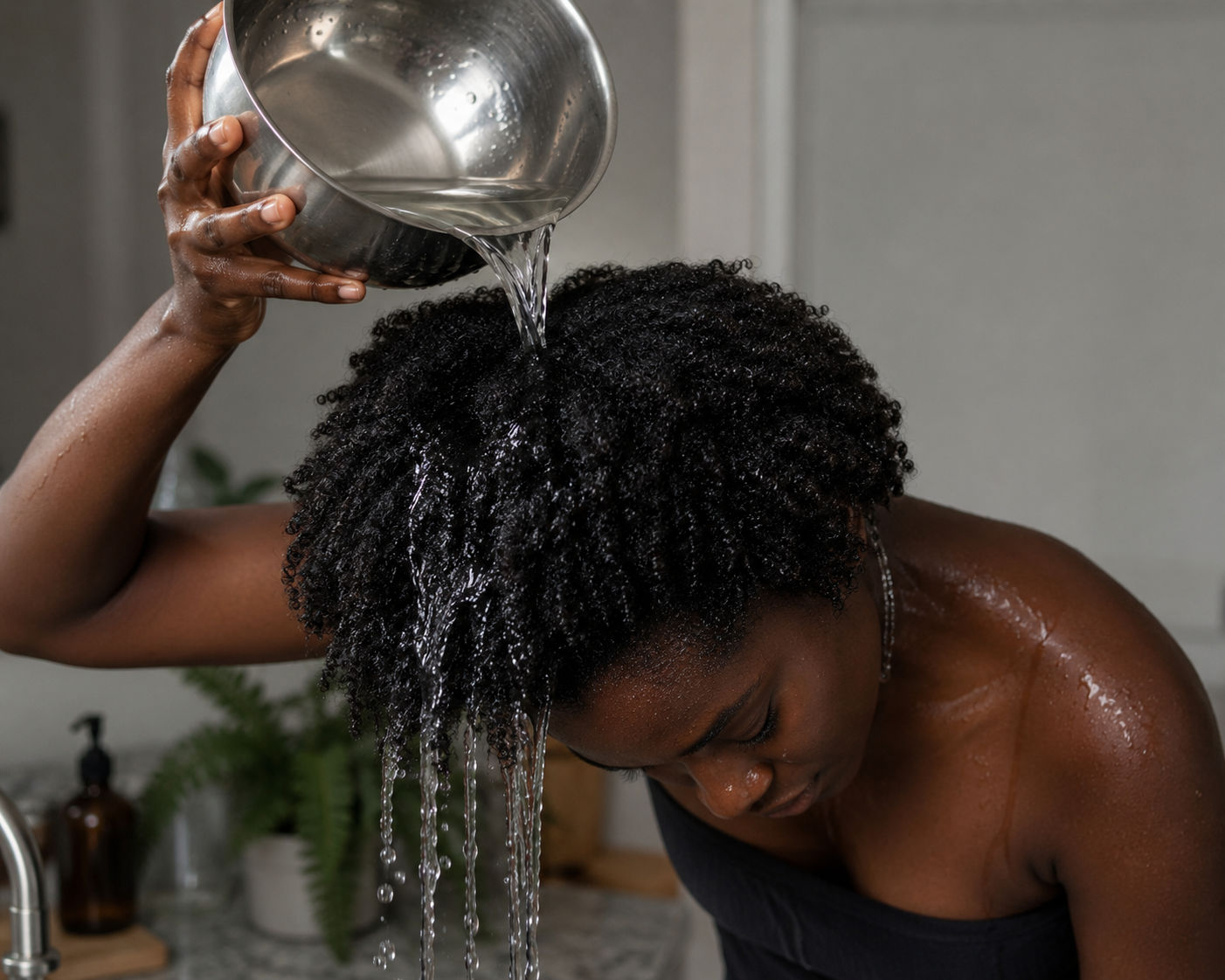 Distilled water being poured over natural hair from a metal bowl, held over a sink