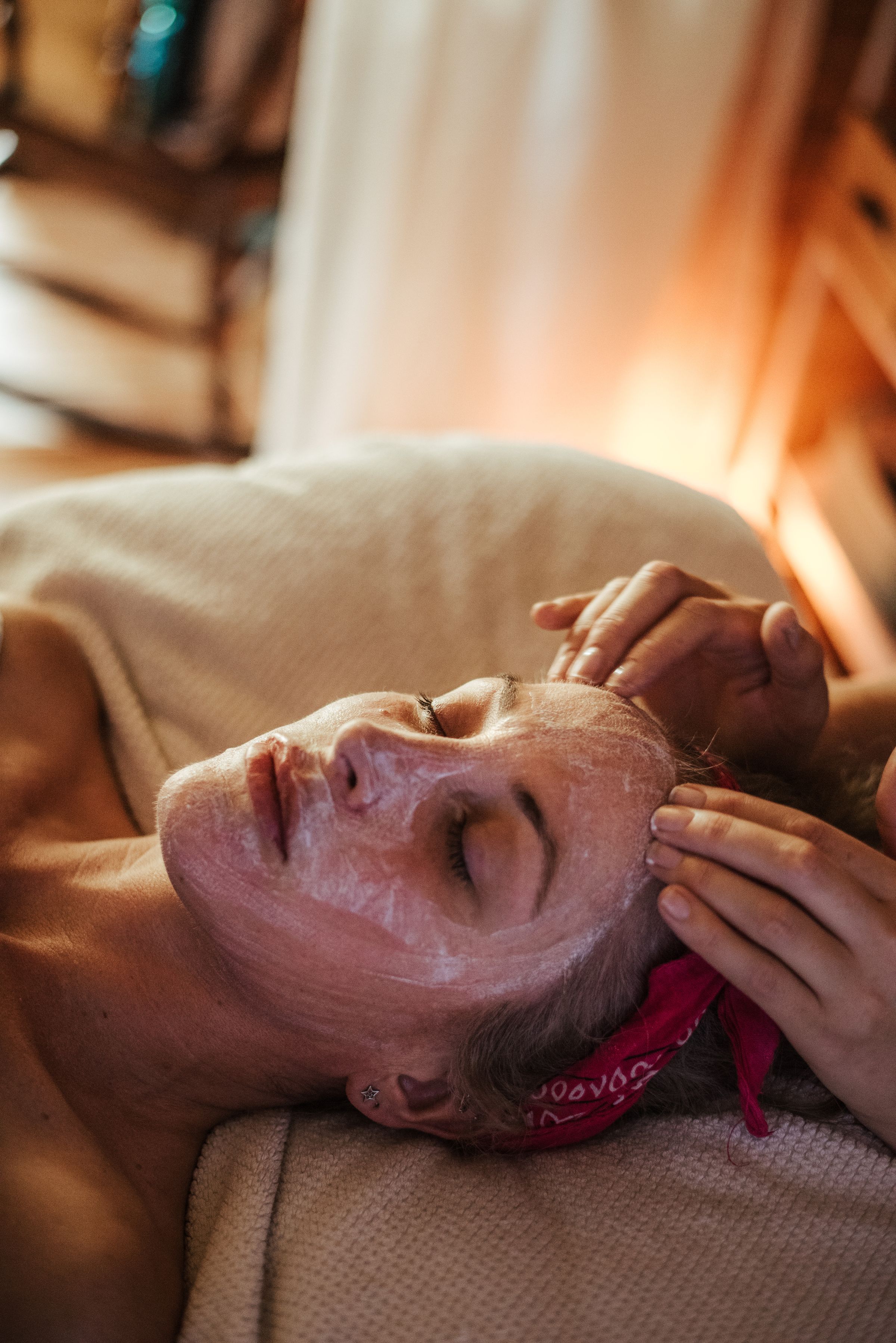 An aesthetician's hands smoothing a treatment mask across a client's face, in warm daylight