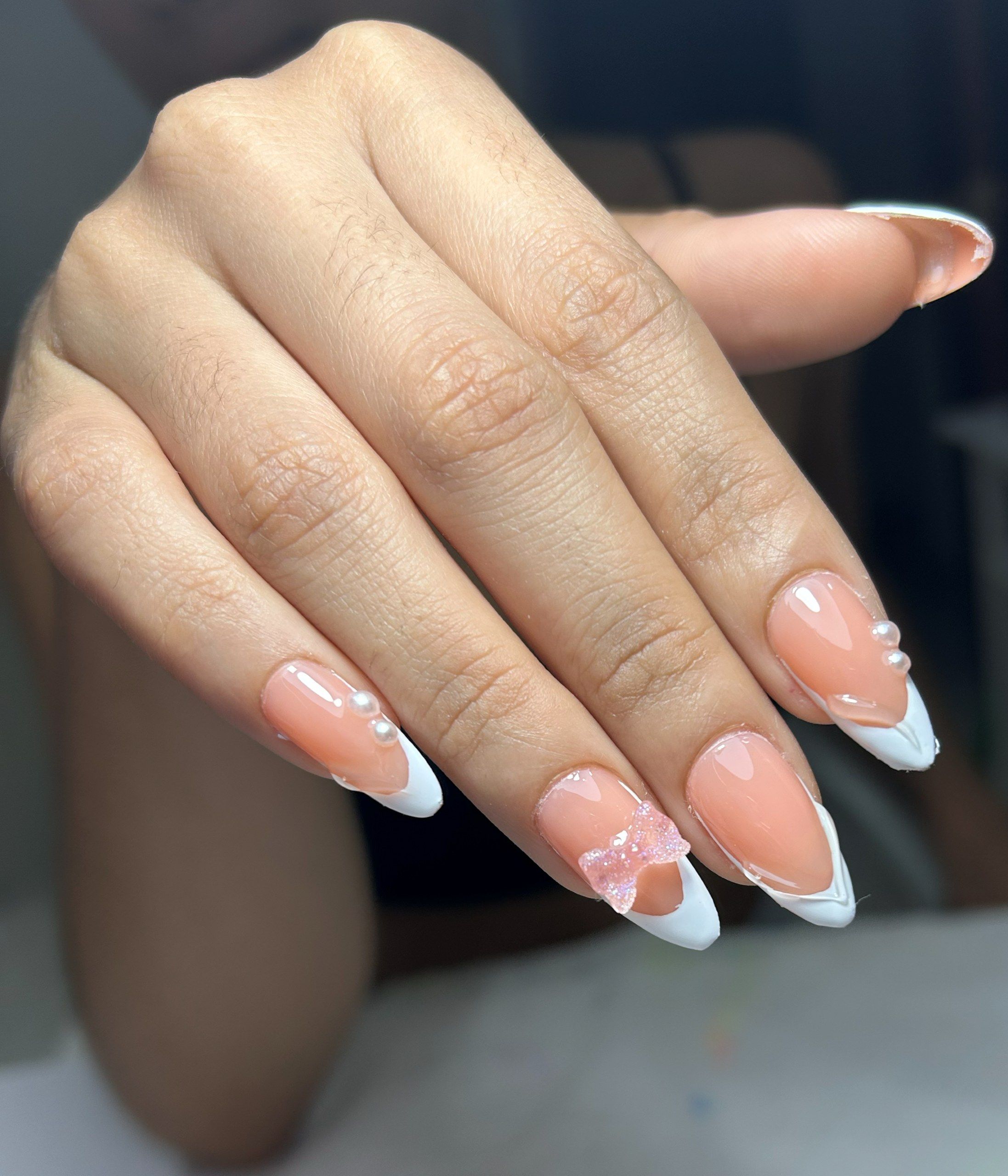 Uñas almendradas con francesa blanca, perlas pequeñas y un moño rosa en relieve en una uña