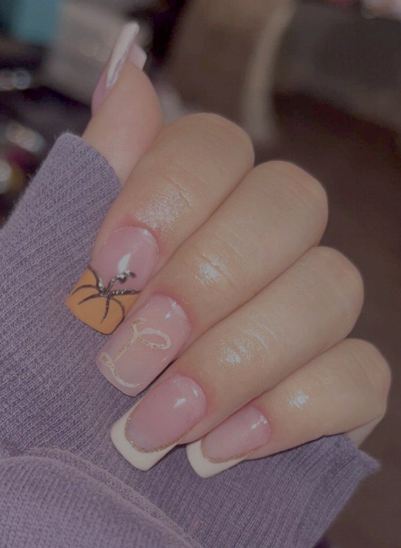 Uñas cuadradas con francesa blanca, una uña naranja con una araña negra pintada y una letra dorada en la siguiente