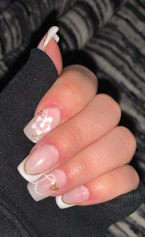Uñas cuadradas con francesa blanca bordeada en dorado, una flor blanca en relieve en una uña y un pequeño dije dorado en otra