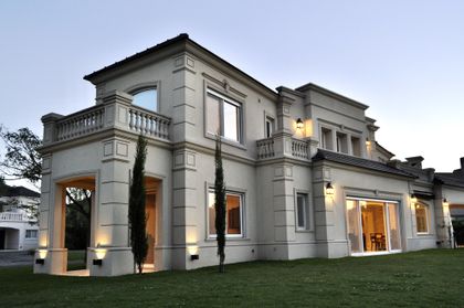 Casa clasica gris con fachada iluminada al atardecer, jardin frontal — proyecto Ayres del Pilar, por Marconi Silva Arquitectos, en Pilar