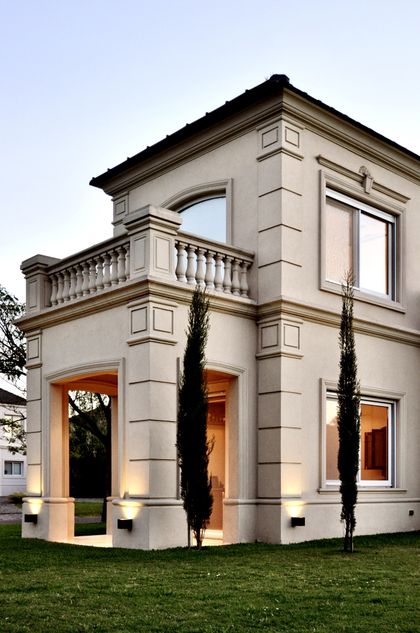 Casa moderna de fachada blanca con balcón y cipreses iluminada — proyecto Ayres del Pilar, por Marconi Silva Arquitectos, en Pilar