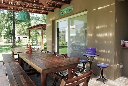 Galería techada con mesa de madera y vista al jardín verde — proyecto El Encuentro  2, por Marconi Silva Arquitectos, en Benavidez