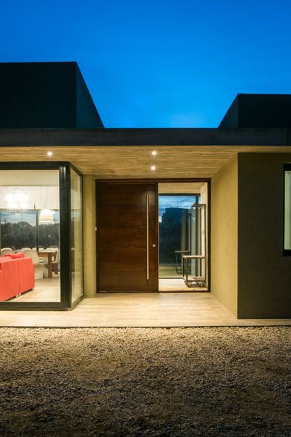 Fachada moderna con puerta de madera, ventanales y grava al atardecer — proyecto Costa Esmeralda 3, por Arquitecto Caceres, en Costa Esmeralda