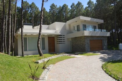 Fachada moderna blanca con detalles de madera en entorno boscoso — proyecto PINAMAR NORTE, por LADAGA-RUARTE-Pinamar, en Pinamar