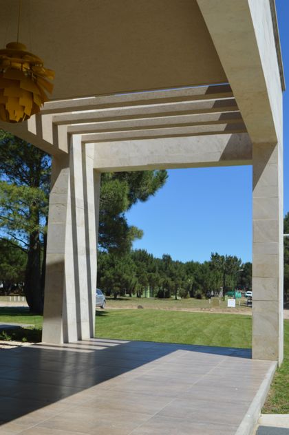 Pérgola moderna con luces colgantes amarillas y césped al fondo — proyecto LA HERRADURA - Boulevard III, por LADAGA-RUARTE-Pinamar, en Pinamar