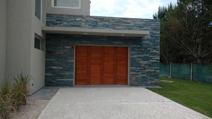 fachada moderna con revestimiento de piedra y portón de madera — proyecto LA HERRADURA - Cul de Sac De La Cincha, por LADAGA-RUARTE-Pinamar, en Pinamar
