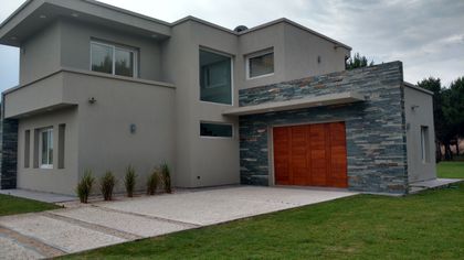 Fachada moderna de casa con detalles en piedra y puerta de garaje de madera — proyecto LA HERRADURA - Cul de Sac De La Cincha, por LADAGA-RUARTE-Pinamar, en Pinamar