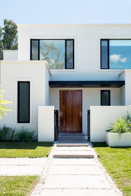 Fachada moderna blanca con puerta de madera oscura y camino de hormigón — proyecto Bosque Chico, por Estudio Arq. María Sioli, en Garin