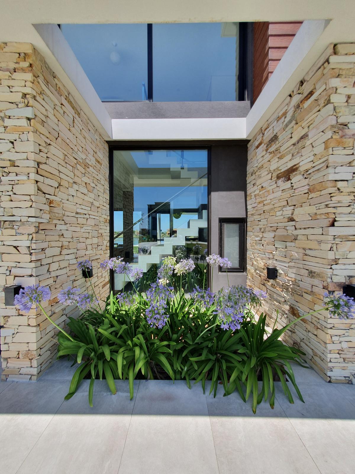Fachada moderna com pedra empilhada, janelas altas e jardim floral — projeto CASA PUERTOS, por ABAD GADANO, em Belén de Escobar