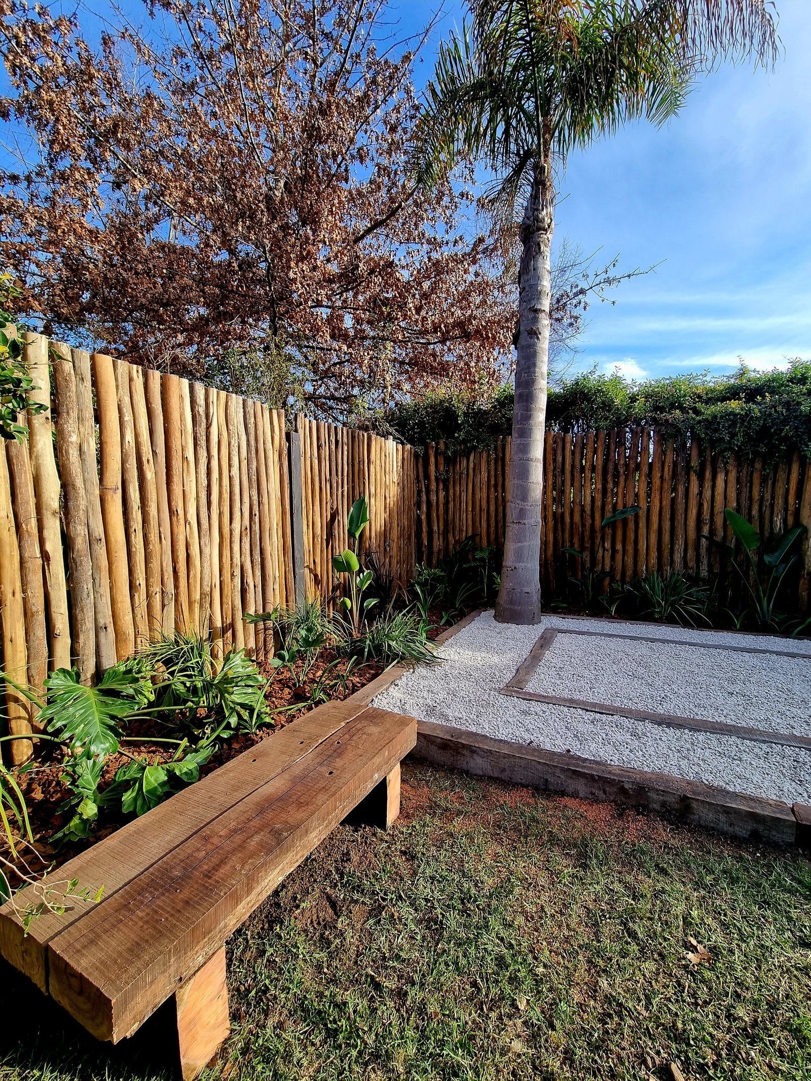 Banquito rústico de madera en jardín con palmera — proyecto Barrio La Rinconada - Pilar, por Marcela García Brünner Paisajista, en La Rinconada