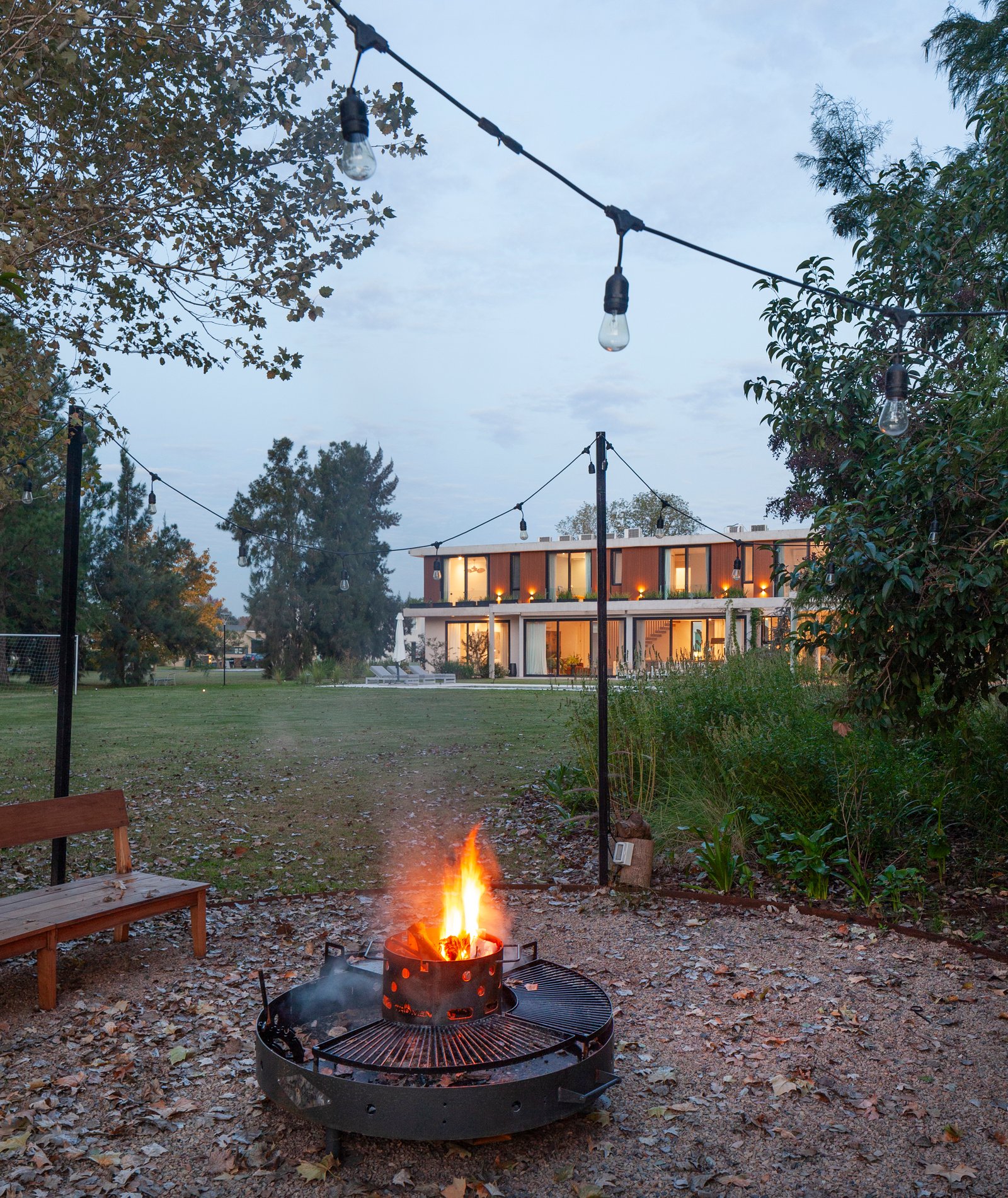 Pátio moderno com fogueira e luzes suspensas em frente à casa — projeto CASA HS143, por A'PRIMA - Arquitectura Sustentable, em km 7.5 Ruta 25
