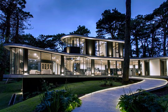 Casa moderna con fachada curva de vidrio en bosque nocturno — proyecto Casa Ameba, por MUCHTEK, en Cariló