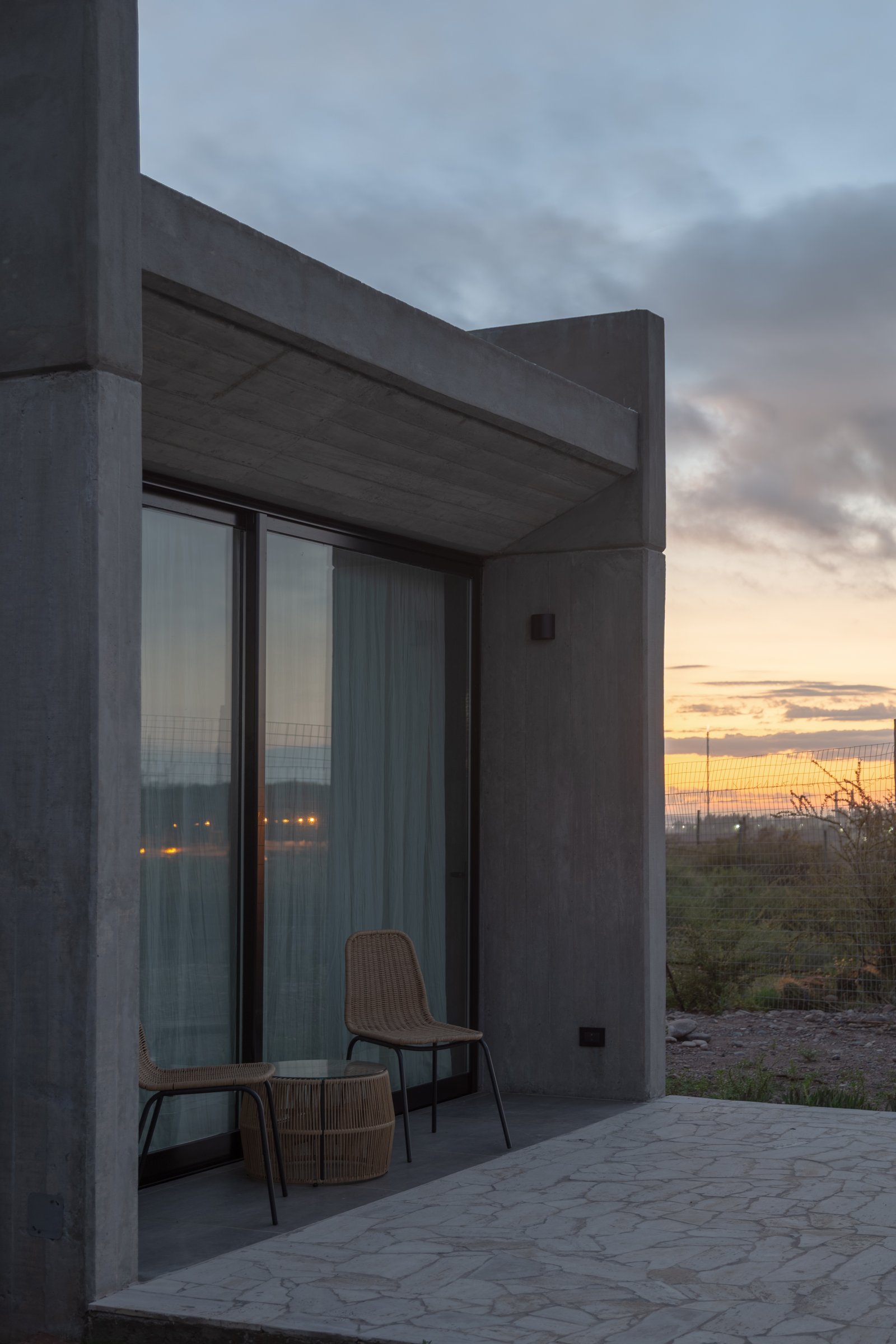 Terraza de hormigón visto con sillas de rattan al atardecer — proyecto Complejo SCV, por ONA - Oficina Nómada de Arquitectura, en Las Compuertas