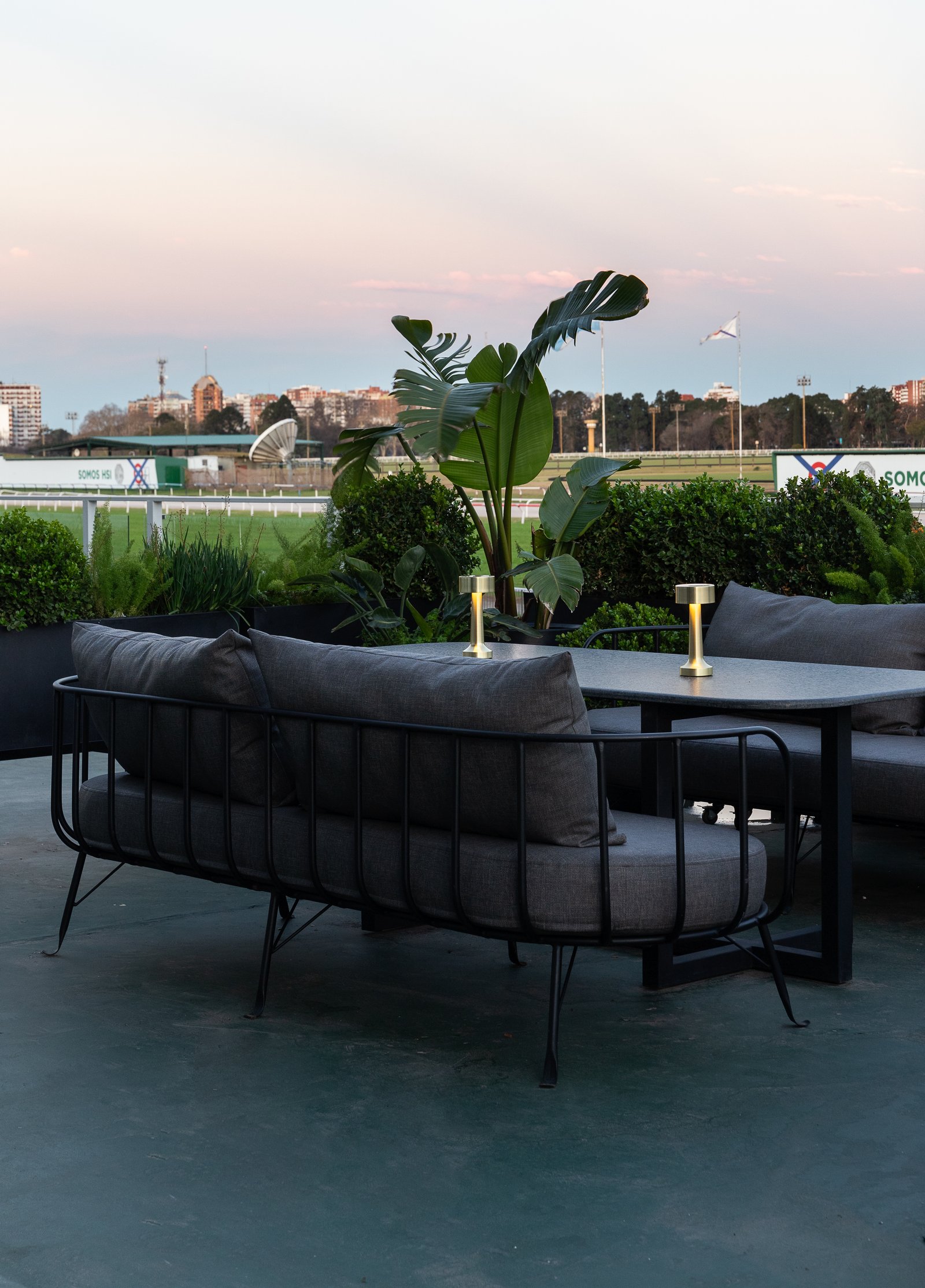 Sofá moderno externo cinza com mesa e plantas em frente a uma pista — projeto PRESIDENTE BAR HIPODROMO, por AB ARQUITECTURA, em San Isidro