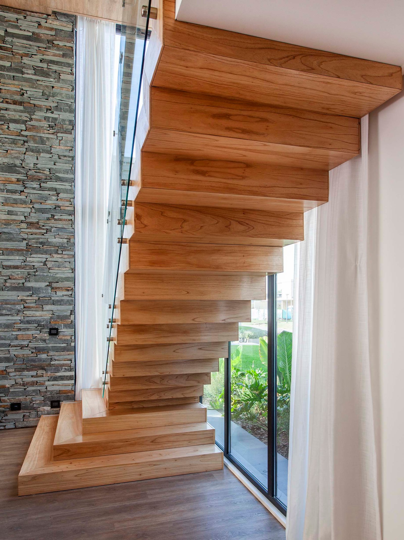 Escalera flotante moderna de madera con pared de piedra y ventanal — proyecto CASA T199, por A'PRIMA - Arquitectura Sustentable, en Los Robles 5901