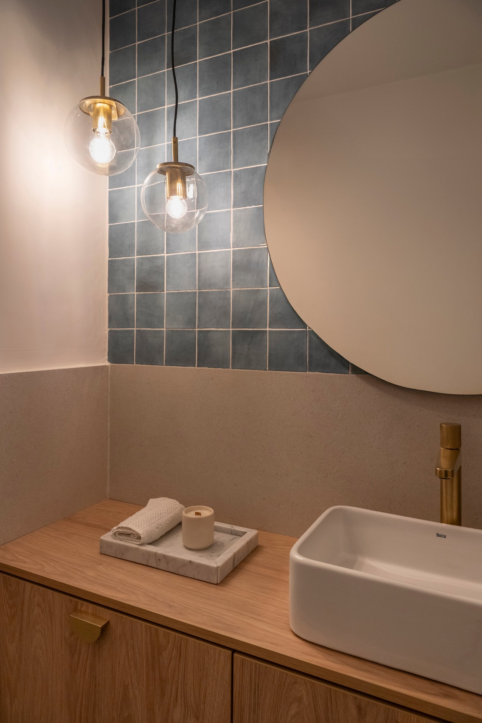 Baño moderno con pared de azulejos azules, espejo circular y vanitory de madera — proyecto Ballester, por BK Arquitectos, en B1653 Villa Ballester
