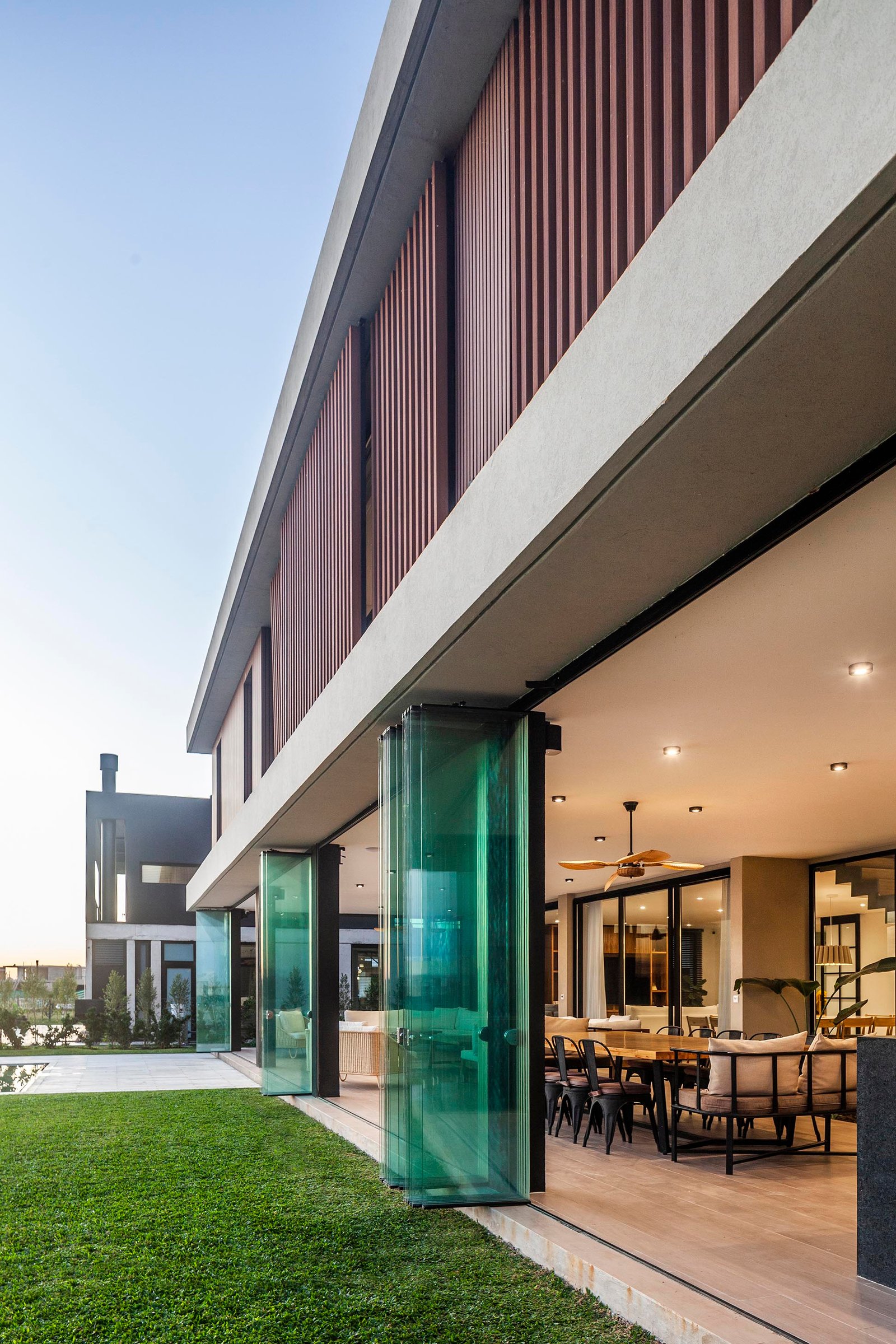 Fachada moderna con puertas de vidrio verde y muro de lamas de madera — proyecto CASA C313, por A'PRIMA - Arquitectura Sustentable, en Provincia de Buenos Aires