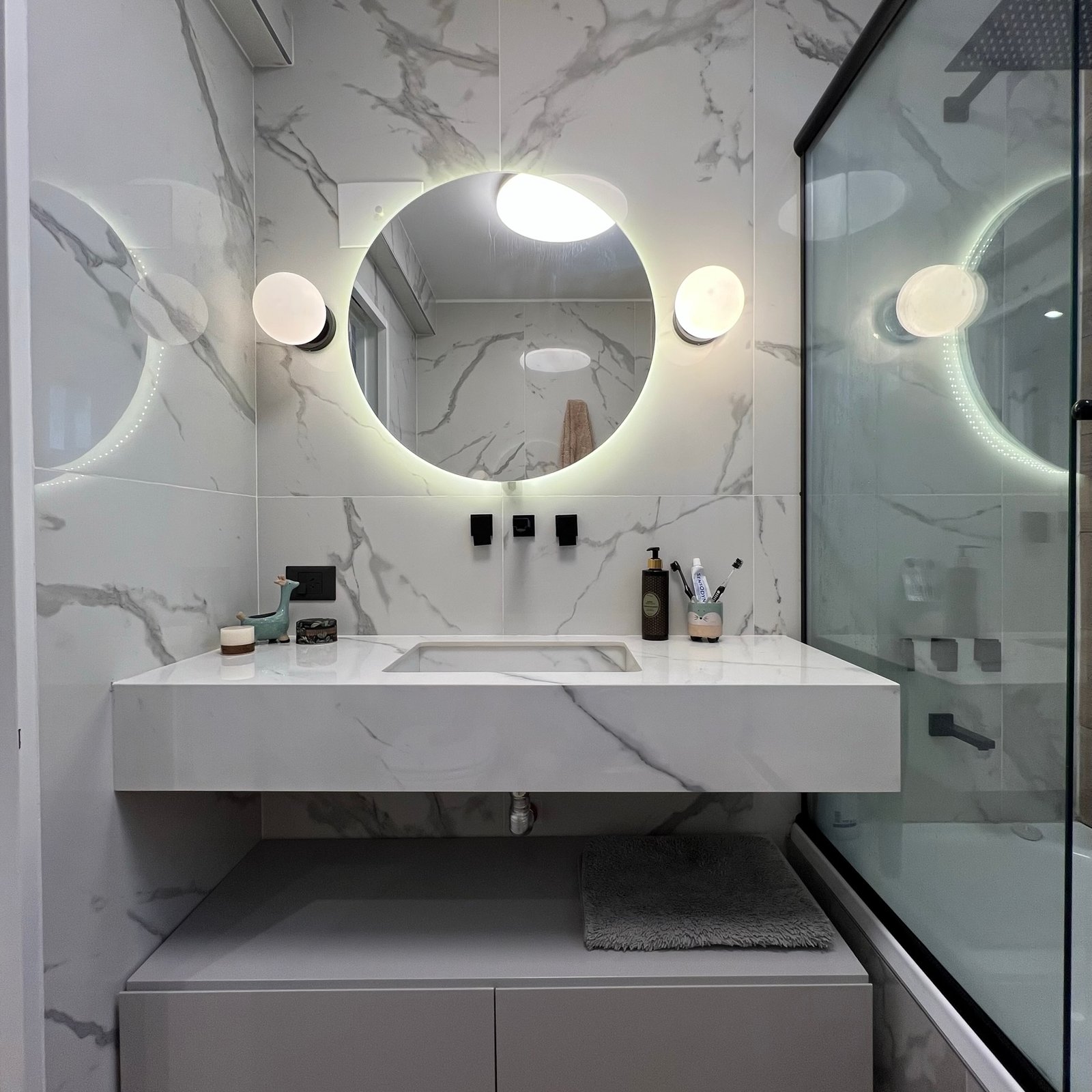 Baño moderno con mármol blanco, vanitory gris y espejo circular retroiluminado — proyecto Diaz Velez I, por MDBK Arquitectura, en Almagro