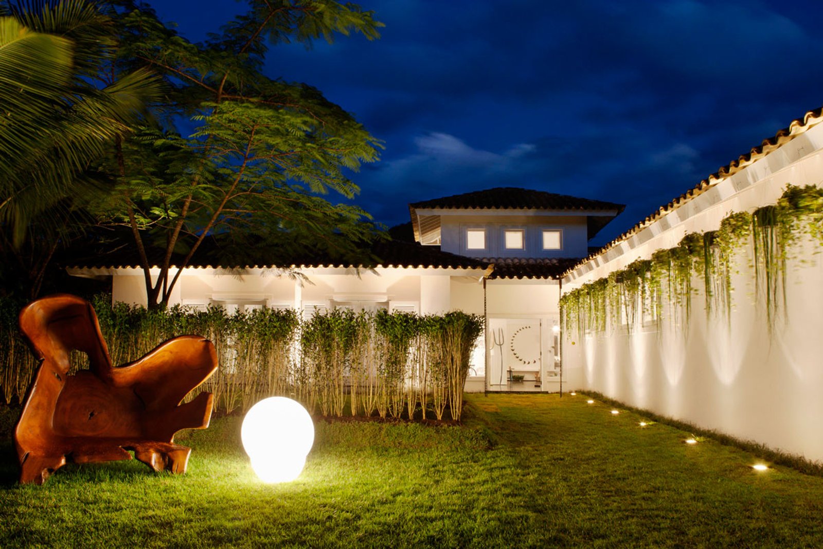 Patio nocturno con iluminación, escultura naranja y jardín vertical — proyecto Arquitetura e Interiores I, por Demian Golovaty