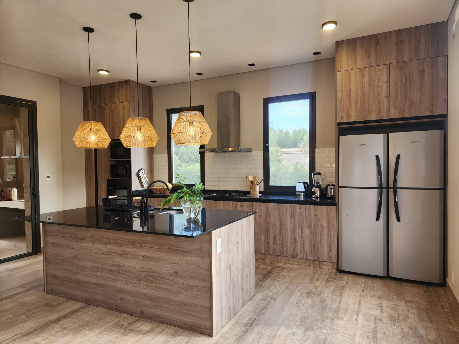 Cocina moderna de madera natural con isla grande y luces colgantes negras — proyecto Ca La Estancia, por Luciana González Anderson, en Cafayate