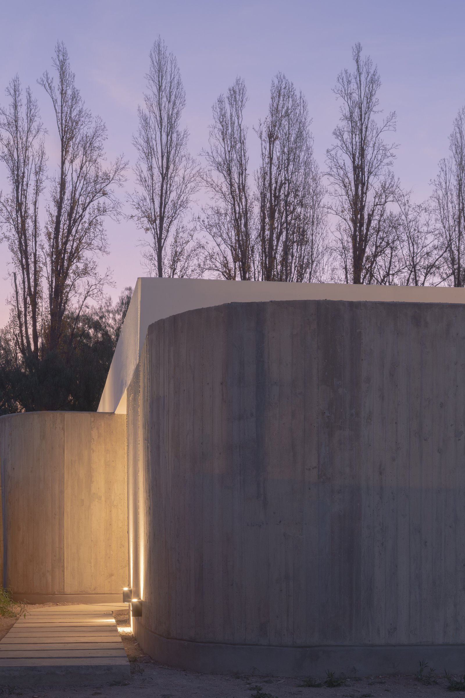 fachada moderna con muros curvos de hormigón y árboles altos al atardecer — proyecto Vivienda CCE, por ONA - Oficina Nómada de Arquitectura, en M5505 Chacras de Coria