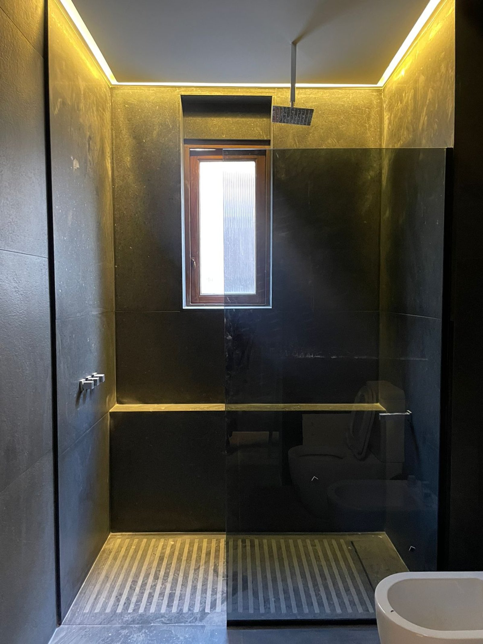 Baño moderno con iluminación cálida, paredes de piedra y mampara de vidrio — proyecto Uruguay y Juncal , por Estudio Babilonia , en Buenos Aires