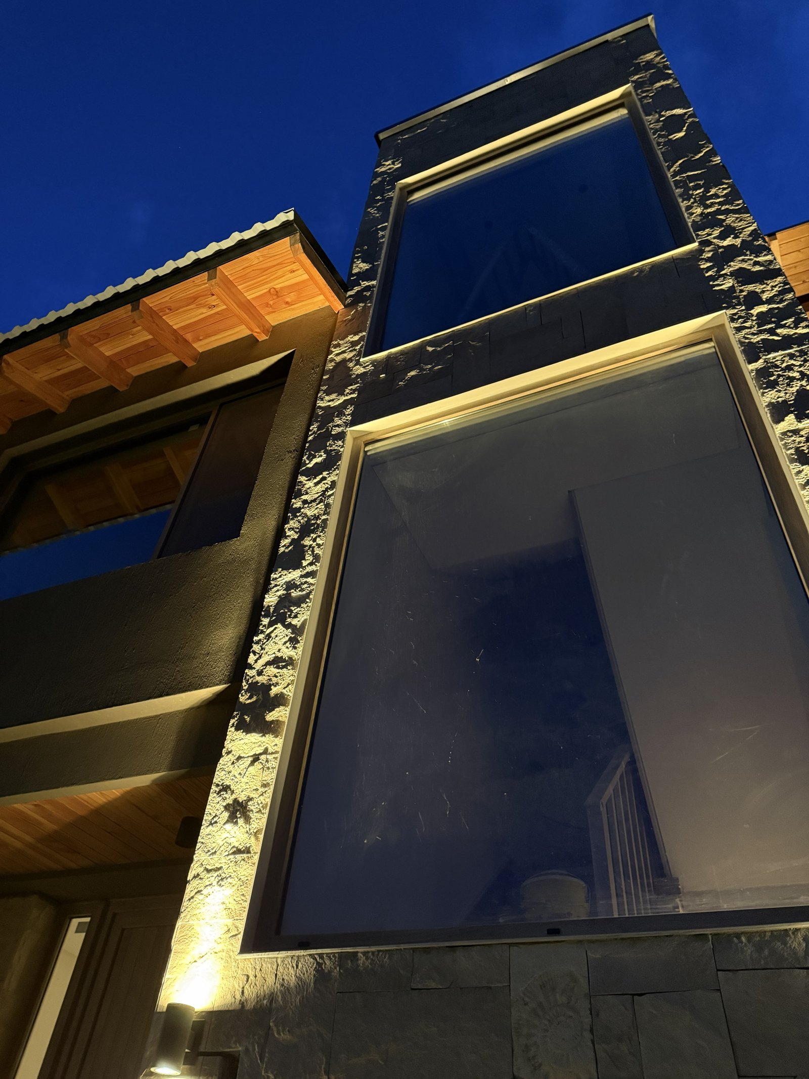 Fachada moderna con grandes ventanales y detalles en piedra, iluminada cálidamente por la noche — proyecto Barrancas de Melipal, por Arquitecta Grace Overgaard, en Av. Ezequiel Bus