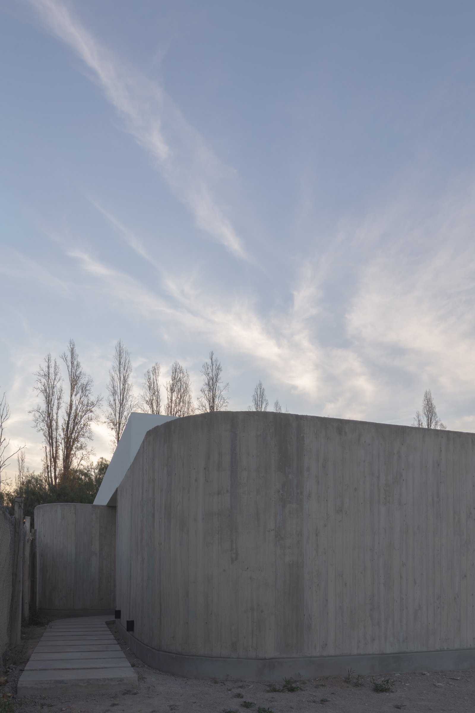 Fachada de concreto moderno con muro curvo y árboles esbeltos — proyecto Vivienda CCE, por ONA - Oficina Nómada de Arquitectura, en M5505 Chacras de Coria