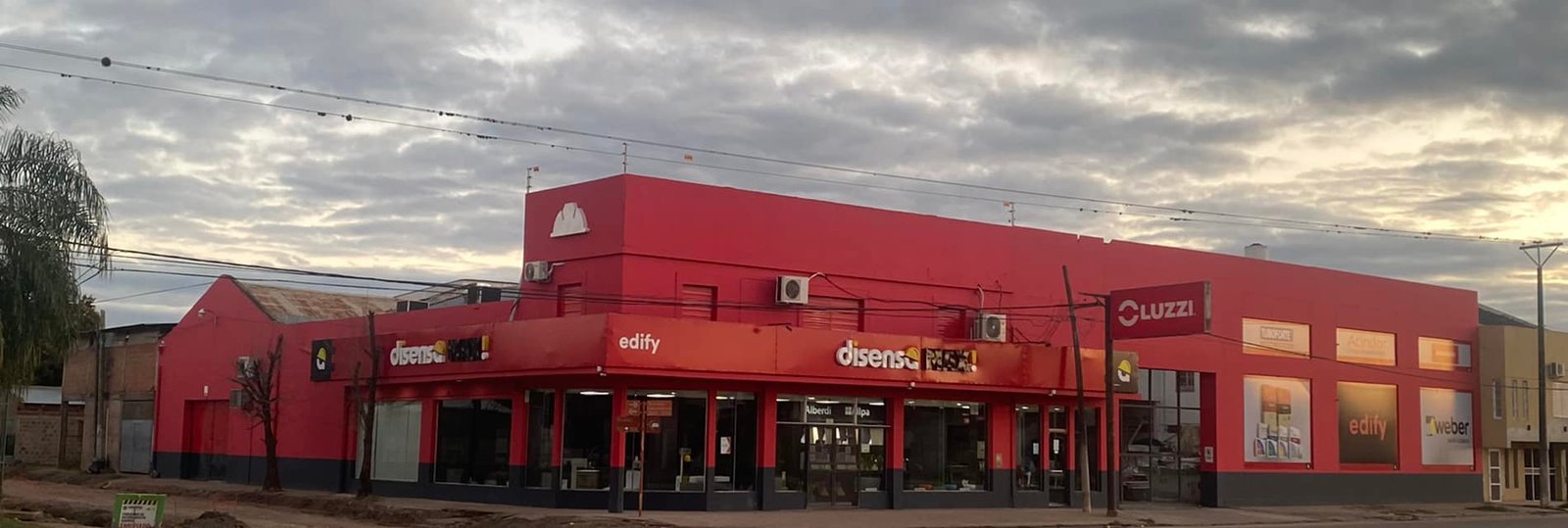 Fachada vermelha de loja comercial com toldo laranja e letreiros — projeto Edify Casa central. Local de venta de materiales de Construccion., por Arquitecto Gustavo Maidana, em Cor